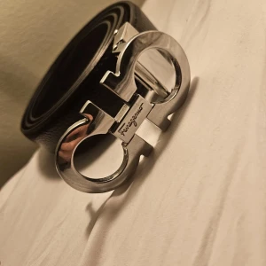 Ferragamo bälte - Säljer ett svart skärp från Ferragamo med ikoniskt dubbel-Gancini-spänne i blankt silver. Bältet är gjort i äkta skinn och har en stilren, klassisk design som lyfter vilken outfit som helst. Perfekt för dig som gillar exklusiva accessoarer.