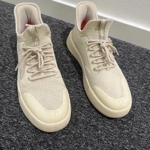 Beige sneakers från C'est Normal - Snygga beige sneakers från C'est Normal med stickad ovandel och robust sula i vitt och orange. Skorna har snörning, rund tå och coola detaljer som dragflik bak och märkning på sulan. Perfekta för dig som gillar stilrena och moderna sneakers.