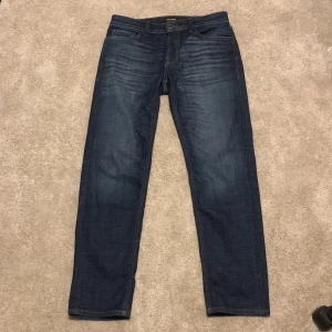 Jack & Jones mörkblå tapered jeans - Mörkblå jeans från Jack & Jones med klassisk femficksdesign och snygga slitningar framtill. Storlek 31/32. Modellen heter Tapered/Mike och har en avsmalnande passform som sitter skönt över benen. Tillverkade i slitstarkt bomullsmaterial med läderpatch bak i midjan.