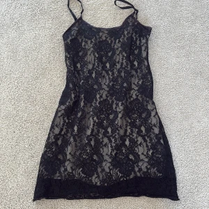 Svart spetsklänning från Impulz - Säljer en svart kortklänning från Impulz i storlek S. Klänningen har tunna axelband, dragkedja i ryggen och är täckt av ett blommigt spetsmönster över ett beige foder. Perfekt för dig som gillar en elegant och edgy stil.