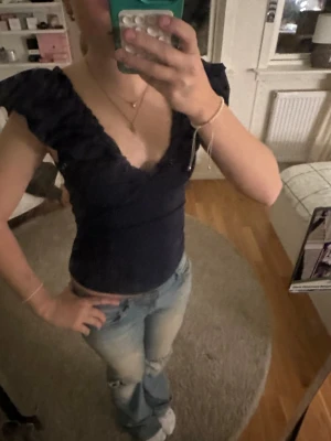 Mörkblå topp med volang från Holly & Whyte - Supersnygg mörkblå topp från Holly & Whyte med volangdetaljer längs den djupa v-ringningen och axlarna. Modellen är tight och ärmlös, perfekt för att lyfta din outfit. Materialet är mjukt och stretchigt polyester som sitter skönt mot kroppen.