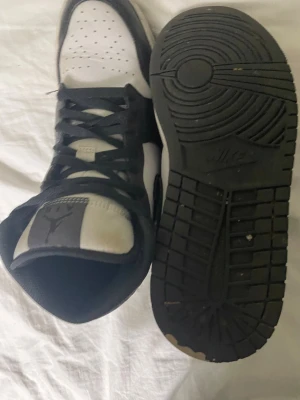 Nike Air Jordan 1 svart/vit sneakers - Säljer ett par Nike Air Jordan 1 sneakers i svart och vitt med klassisk high-top design. Skorna har snörning, perforerad tå och rund tå. Materialet är skinn och de har en platt sula. Perfekt för dig som gillar streetwear och vill ha en ikonisk look.