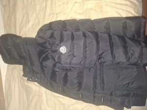 Svart Moncler dunjacka med huva - Svart dunjacka från Moncler med huva och klassisk Moncler-logga på bröstet. Jackan har quiltad design, dragkedja framtill och praktiska fickor. Insidan har en unik serietidningsetikett. Perfekt för kalla dagar och riktigt snygg streetstil. Jackan kommer att levereras med prislapparna kvar på den.