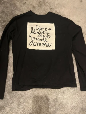 Svart långärmad tröja Zara med tryck - Svart långärmad tröja från Zara med ett stort vitt tryck på ryggen där det står 'Ciao e benvenuto grande amore' i handskriven stil. Tröjan är i mjuk bomull och har en avslappnad passform, perfekt för dig som gillar coola statement-plagg.