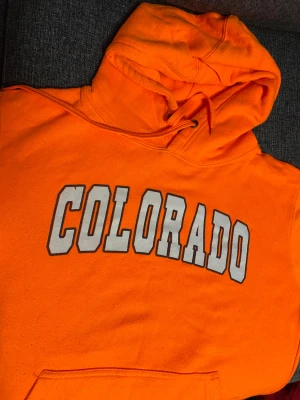 Orange Colorado hoodie från EAGLE - Snygg orange hoodie från EAGLE med stor 'COLORADO'-text i vitt och grått på bröstet. Tröjan har huva med snörning och en klassisk magficka. Tillverkad i mjuk bomull och polyester, perfekt för dig som gillar färgstarka plagg.