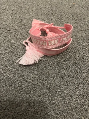 Christian Dior rosa armband med tofsar - Säljer ett par snygga rosa armband från Christian Dior med broderad text och dekorativa tofsar i ändarna. Armbanden har justerbar knytning och är tillverkade i ett mjukt tygmaterial. Perfekt accessoar för att ge din outfit en lyxig touch.