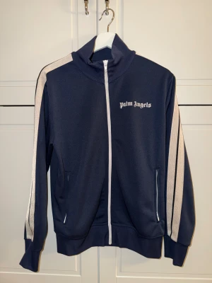 Navy / Cream Palm Angels - Snygg marinblå kofta från Palm Angels med vita ränder längs ärmarna och vit dragkedja framtill. Jackan har hög krage, två fickor med dragkedja och logotypen tryckt på bröstet. Perfekt för en sportig och trendig look.