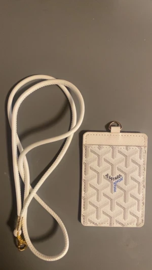 Vit plånbok med snodd - Snygg vit plånbok med klassiskt geometriskt mönster och blå logga. Kommer med matchande vit snodd och guldfärgad hake, perfekt för att bära runt halsen. Fönster på baksidan för ID eller kort. Stilren och praktisk accessoar.