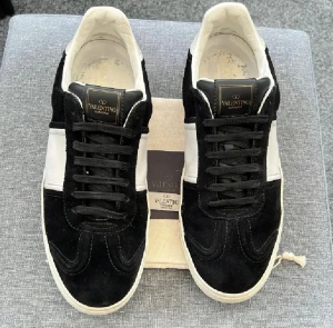 Valentino flycrew  - Svarta och vita sneakers från Valentino Garavani med snygg kontrastpanel i vitt på sidan. Skorna har rund tå, platt sula och är tillverkade i mocka och skinn. Klassisk snörning och diskret logga på plösen ger en stilren vibe. alla borrar kvar 