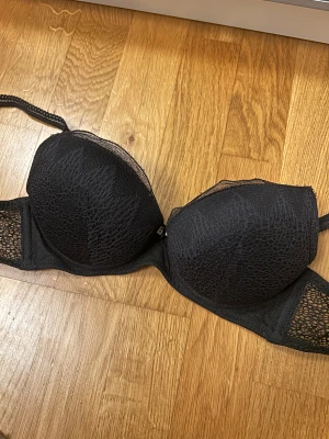 Svart spets bh Intimissimi - Supersnygg svart bhfrån Intimissimi med snygga spetsdetaljer och meshpartier. Kuporna har ett diskret mönster och en liten rosett framtill. Storlek 70b och knappt använd!