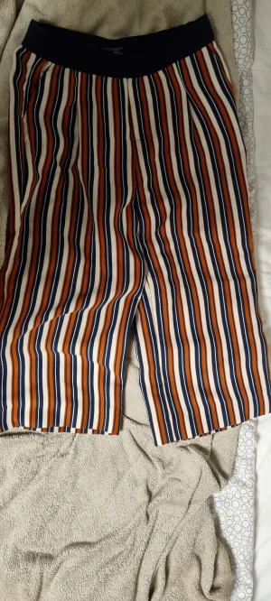 Randiga byxor från Zara Trafaluc - Snygga randiga byxor från Zara Trafaluc med elastisk midja och raka ben. Färgerna är marinblå, orange och vit, vilket ger en cool och ungdomlig vibe. Materialet känns lätt och skönt, perfekt för varmare dagar. Bakfickor med dekorativa detaljer.