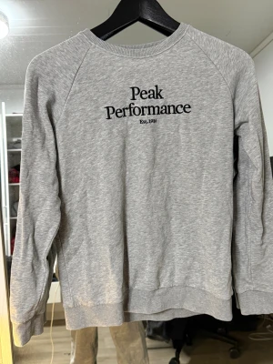 Grå Peak Performance sweatshirt - Snygg grå sweatshirt från Peak Performance med svart broderad logga på bröstet. Tröjan har rund hals, ribbade muddar och är tillverkad i mjuk bomullsmix. Perfekt för chill dagar och enkel att matcha med jeans eller joggers.