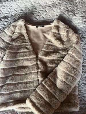 Beige pälsjacka från Gina Tricot - Supermjuk och fluffig beige pälsjacka från Gina Tricot med horisontella ränder i materialet. Jackan har lång ärm och öppen front, perfekt för dig som gillar en cozy och trendig look. Fodrad insida för extra komfort.
