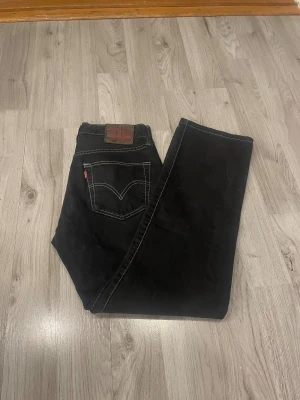 Svarta Levi's 501 jeans med kontrastsömmar - Svarta Levi's 501 jeans med klassisk rak passform och tydliga vita kontrastsömmar. Jeansen har det ikoniska läderpatchen bak och röd Levi's-tag på bakfickan. Materialet är robust denim och modellen har fem fickor.
