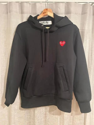Ej till salu  - Svart hoodie från Comme des Garçons Play med rött hjärta med ögon broderat på bröstet. Tröjan har huva med snörning, långa ärmar och praktisk magficka. Perfekt för dig som gillar stilrena streetwear-plagg med en twist.