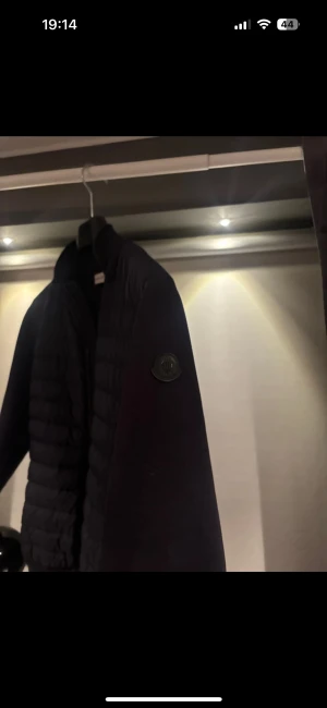 Svart dunjacka från Moncler - Svart dunjacka från Moncler med quiltad framsida och mjuka ärmar. Jackan har lång ärm, ribbade muddar och Moncler-logga på armen. Materialet är en mix av dun och polyester, perfekt för kyliga dagar. Snygg och stilren design som lyfter din outfit. Den är köpt på NK för 4 år sedan. 