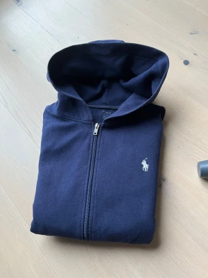 Mörkblå hoodie från Polo Ralph Lauren. Reserverad! - Säljer en mörkblå hoodie från Polo Ralph Lauren med dragkedja och klassisk logga på bröstet. Tröjan har huva, fickor framtill och är tillverkad i mjuk bomull. Perfekt för chill dagar och enkel att matcha med jeans eller joggers.