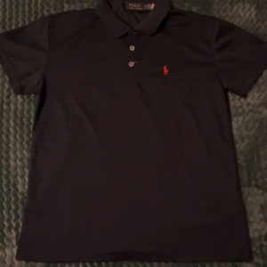 Blå  pikétröja från Ralph Lauren - Klassisk svart pikétröja från Ralph Lauren med röd broderad logga på bröstet. Tröjan har korta ärmar, krage och knäppning med tre knappar framtill. Perfekt för en stilren och sportig look.