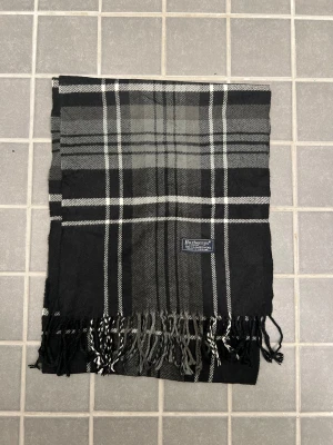 Svart och grå rutig halsduk Burberrys - Klassisk rutig halsduk från Burberrys i 100% ull. Halsduken har fransar i ändarna och den ikoniska Burberrys-etiketten. Perfekt accessoar för att lyfta din outfit. Anledningen att de stör ”burberrys” är för att de är en äldre modell.