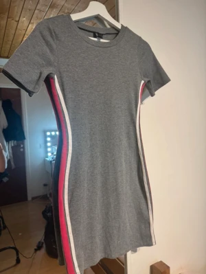 Grå kortklänning med ränder - Grå kortklänning med tight passform. Klänningen har vita och röda ränder längs sidorna. Super bra skick! 
