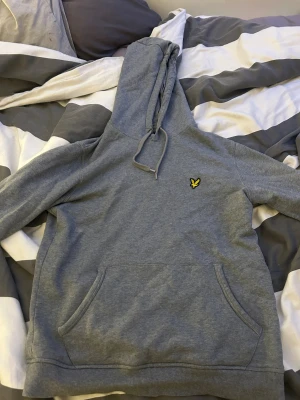 Grå hoodie från Lyle & Scott - Grå hoodie från Lyle & Scott med klassisk gul fågel-logga på bröstet. Tröjan har huva med snörning, stor magficka och långa ärmar. Mjuk bomullskänsla och relaxed fit, perfekt för chill dagar. Enkel och stilren design som funkar året runt, dock en liten fläck på ryggen.