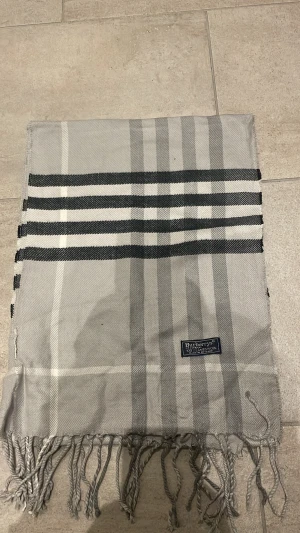 Grå rutig halsduk från Burberry - Stilren grå halsduk från Burberrys i klassiskt rutigt mönster med svarta och vita detaljer. Halsduken är tillverkad i 100% lammull och har tvinnade fransar i kanterna. Perfekt accessoar för att lyfta din outfit.