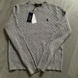Grå stickad Ralph lauren tröja - Snygg grå kabelstickad tröja från Polo Ralph Lauren med klassisk rund hals och svart broderad logga på bröstet. Tröjan har långa ärmar och ribbade muddar vid ärmslut och nederkant. Perfekt för dig som gillar stilrena och tidlösa plagg.