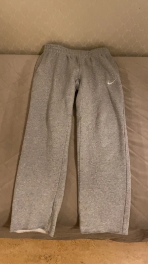 Grå mjukisbyxor från Nike - Grå mjukisbyxor från Nike med klassisk vit swoosh-logga på vänster ben. Byxorna har elastisk midja, raka ben och är tillverkade i mjukt bomullsmaterial. Perfekta för chill eller träning, med fickor på sidorna och enkel design. De är flared och klippna.