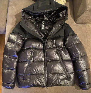 Svart pufferjacket från Replay - Svart jacka från Replay med glansig yta. Jackan har två fickor med dragkedja samt quiltad design. Väldigt varm så perfekt för kalla dagar. Inga skador eller lagningar någonstans på jackan.