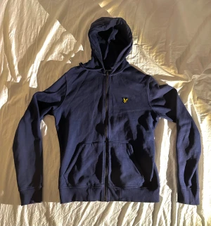 Marinblå hoodie från Lyle & Scott - Snygg marinblå hoodie från Lyle & Scott med gul logga på bröstet. Tröjan har huva med snörning, dragkedja framtill och två stora fickor. Tillverkad i mjuk bomull, perfekt för chill dagar. Klassisk passform och långa ärmar.