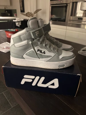 Fila gråvita high-top sneakers strl 42 - Säljer ett par Fila high-top sneakers i grått och vitt med snygga detaljer och logga på sidan och hälen. Skorna har snörning, kardborreband och platt sula. Tillverkade i syntetläder med textilfoder och gummisula. Perfekt för dig som gillar sportig och clean stil.