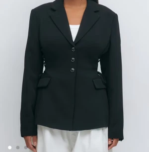 Svart kavaj med snörning i midjan - Stilren svart kavaj med tre knappar framtill och snygga ficklock. Kavajen har unik snörning i midjan som ger en cool och figurnära siluett. Klassisk krage och långa ärmar. Perfekt för dig som vill ha en modern twist på en klassiker. Helt slutsålt på hemsidan 