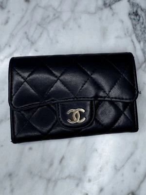 Svart quiltad Chanel plånbok i skinn - Add a touch of effortless luxury to your everyday essentials with this iconic Chanel Classic Quilted Card Holder. Crafted in soft black lambskin with Chanel’s signature diamond quilting, it’s finished with the timeless gold-tone CC logo. • Color: Black • Material: Lambskin leather • Hardware: Gold-tone CC logo • Design: Classic diamond quilting • Function: Card holder / small wallet • Measurements: approx. 7.5 × 11.3 × 2.1 cm 