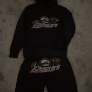 Svart Trapstar hoodie London Shooters - Svart hoodie från Trapstar med stort broderat London Shooters-tryck och pantermotiv på bröstet. Klassisk huva med snörning och ribbade muddar. Perfekt streetwear-vibe och riktigt snygg detalj med kontrastfärger i trycket. Det är XS men passar mer åt S