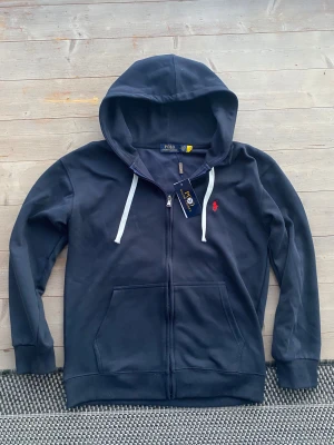 Mörkblå hoodie från Polo Ralph Lauren - Snygg mörkblå hoodie från Polo Ralph Lauren med vit snörning och dragkedja framtill. Tröjan har huva och det klassiska röda Polo-logot broderat på bröstet. perfekt för chill dagar. Fickor på sidorna och ribbade muddar.