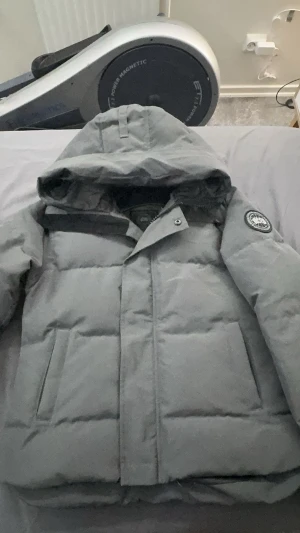 Grå vinterjacka från Canada Goose - Säljer en grå vinterjacka från Canada Goose med huva och klassisk logotyp på ärmen. Jackan har dragkedja och knappar framtill, två sidofickor och quiltad design. Perfekt för kalla dagar med sin tjocka och isolerande dunfyllning. Storlek S.