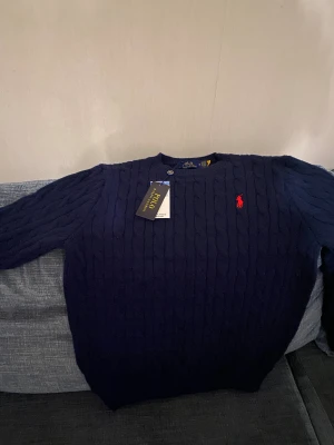 Mörkblå stickad tröja Polo Ralph Lauren - Snygg mörkblå stickad tröja från Polo Ralph Lauren med klassiskt kabelstickat mönster och rund hals. Tröjan har lång ärm och den ikoniska röda Polo-loggan broderad på bröstet. Perfekt för dig som gillar stilrena och tidlösa plagg.