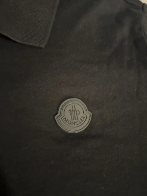 pikétröja från Moncler - Svart pikétröja från Moncler med klassisk krage och två knappar framtill. Tröjan har Moncler-logga i svart på bröstet och är tillverkad i mjuk bomull. Perfekt för dig som gillar stilrena och exklusiva plagg.