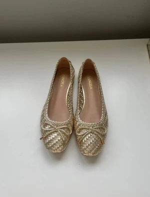 Guldiga ballerinaskor från D'Angela - Snygga guldiga ballerinaskor från D'Angela i flätad syntet med rosett framtill. Skorna har rund tå och platt sula, vilket gör dem både stilrena och bekväma. Perfekta för dig som vill ha en glittrig touch på outfiten.