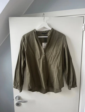 Olivgrön blus med spets från Zara - Olivgrön blus från Zara med snygga spetsdetaljer längs framsidan och ärmarna. Blusen har v-ringning, knappar framtill och långa ärmar med lätt puff vid mudden. Tillverkad i mjuk bomull, perfekt för en trendig och avslappnad look.