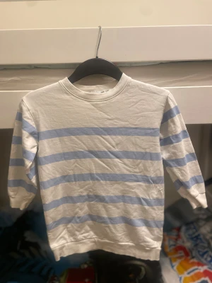 Vit och ljusblå randig tröja 157 - Vit långarmad tröja/kort klänning från 157 med ljusblå horisontella ränder. Rund hals med ribbad kant och mjukt bomullsmaterial. Perfekt för dig som gillar enkel och fräsch stil med lite marint vibe. Barn till tjejer 