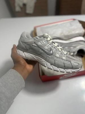 Nike P-6000 grå sneakers - Nike P-6000 sneakers i grått med mesh och syntetpaneler, vita snören och en chunky vit sula. Snyggt sportig design med silverfärgad swoosh på sidan och detaljerade mönster i ovandelen. Perfekt för dig som gillar retrovibe och streetstyle.