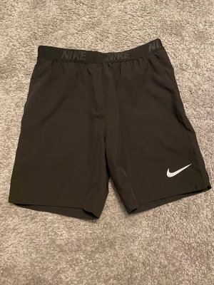 Svarta träningsshorts från Nike - Svarta shorts från Nike med elastisk midja där Nike-loggan är tryckt runt om. Klassisk design med vit swoosh på benet. Perfekta för gymmet eller löpning, tillverkade i lätt polyester som andas och ger skön rörelsefrihet.