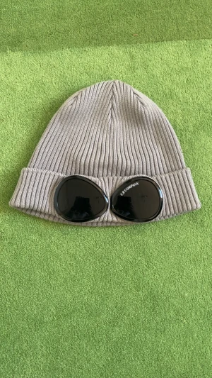 C.P Company Mössa - Snygg grå mössa från C.P. Company med ribbad stickning och coola svarta glasögon-detaljer framtill. Mössan är äkta med Qr kod, finns i Stockholm och kan även fraktas