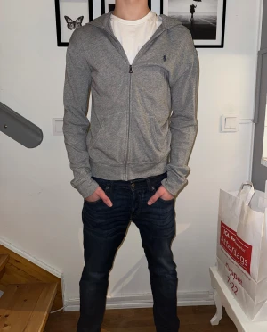 Grå hoodie från Polo Ralph Lauren - Snygg grå hoodie från Polo Ralph Lauren med dragkedja och huva. Tröjan har ett mörkblått broderat Polo-logo på bröstet och är tillverkad i mjuk bomull. Perfekt för chill dagar och har en normal passform med långa ärmar.