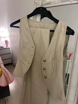 Beige väst med knappar och v-ringning - Stilren beige väst med v-ringning och tre snygga knappar framtill. Västen har figurnära passform och lätt utsvängd nederkant. Tillverkad i ett mjukt tyg som ser ut som bomull eller linne, perfekt för en clean och modern look.