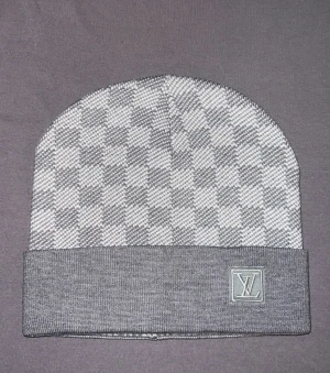 Grå rutig mössa Louis Vuitton - Snygg grå mössa från Louis Vuitton med klassiskt rutigt mönster och uppvikt kant. LV-logga i silver på framsidan ger en lyxig touch. Materialet är mjukt och stickat, perfekt för kyliga dagar. En stilren accessoar som lyfter din look.