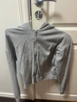 Grå croppad hoodie med dragkedja - Grå hoodie med croppad passform och hel dragkedja framtill. Tröjan har långa ärmar och en stor huva, perfekt för chill dagar. Materialet är mjukt och känns som bomull, vilket gör den skön att bära. Enkel och stilren design utan mönster eller logga.