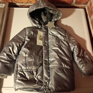 Silver pufferjacka från Bellybutton - Säljer en glansig silverfärgad pufferjacka från Bellybutton med huva och mjukt pälsfoder. Jackan har tryckknappar framtill, ribbade muddar vid ärmslut. Perfekt för kalla dagar och riktigt snygg look. .  Nytt med lappen kvar. Jag kan inte se några hål eller skador etc. Även om de är nytt.  Obs! Jag står inte för frakbolagets slarv, skador eller annat som händer efter att jag har postat.