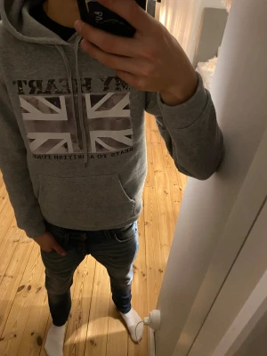 Grå hoodie med brittisk flagga - Grå hoodie med tryck av Union Jack och texten 'MY HEART BEATS TO A BRITISH TUNE' på bröstet. Tröjan har huva med snörning och en stor magficka. Perfekt för dig som gillar streetstyle och vill ha en chill look.